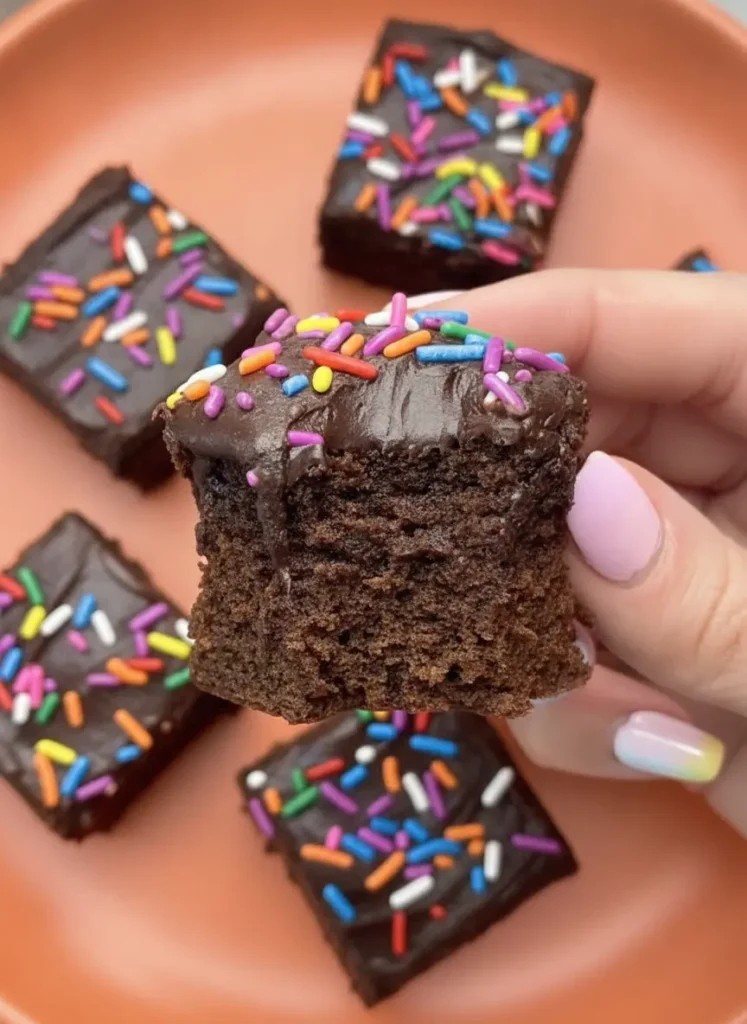 Chocolate Donut-Frosted Cake Bites Recipe - An ingredient prep shot on a wooden cutting board. A small glass bowl of Dutch-process cocoa powder, a ceramic bowl of flour, and a small dish overflowing with vibrant rainbow sprinkles are arranged neatly. A sprig of fresh mint is visible in the background, all captured in natural morning light on a marble surface.