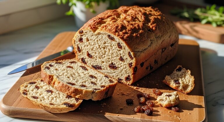 A gloriously baked, golden brown No Knead Cinnamon Raisin Bread loaf, sliced to reveal its tender, raisin-studded crumb, resting on the same wooden cutting board. A few artful crumbs and plump raisins are scattered nearby. The scene is bathed in natural morning light from an east window, highlighting the warm tones of the bread against marble countertops with wood accents. Fresh herbs are visible in the soft-focused background, clean and tidy presentation. No hands.