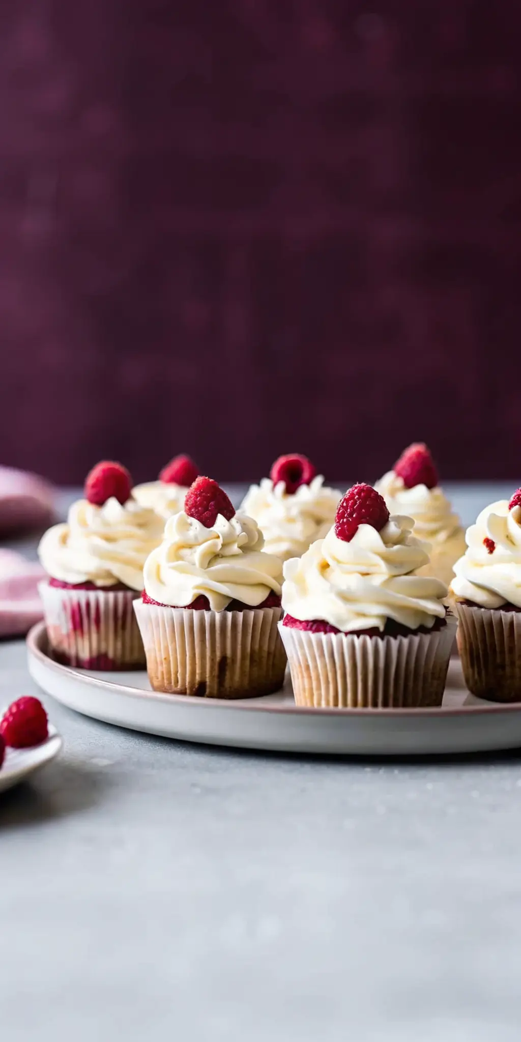 A single White Chocolate Raspberry Cupcake on a small, minimalist white ceramic plate, showcasing the perfectly swirled white chocolate buttercream, glistening fresh raspberries, and delicate white chocolate shavings. The cake liner is light beige, and a peek into the moist, raspberry-studded crumb is visible. Natural morning light, warm tones, soft shadows. 3:4 aspect ratio. NO HANDS OR PEOPLE.