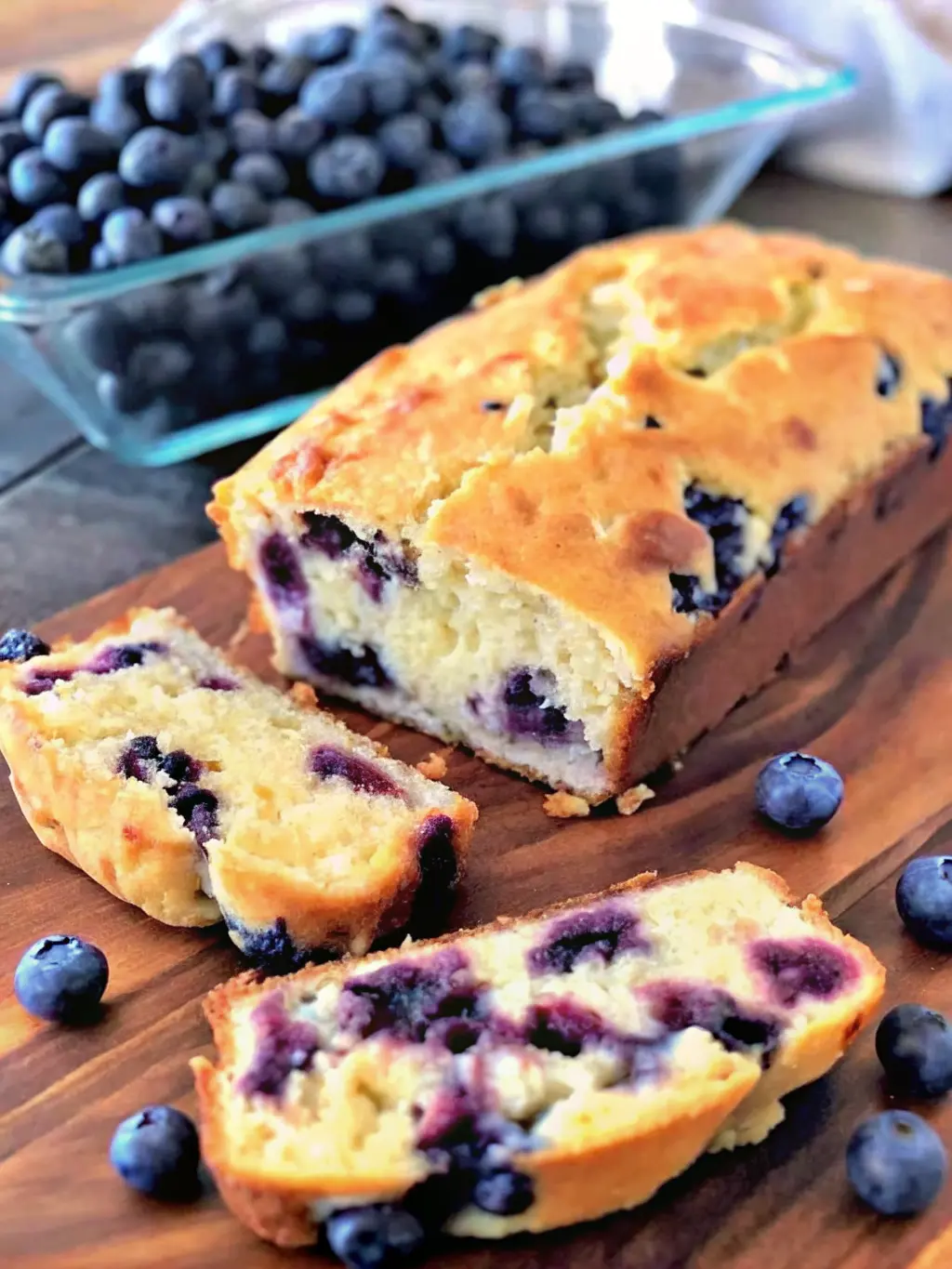A close-up shot of a single, thick slice of Blueberry Cream Cheese Loaf on a minimalist white plate, showcasing the moist, light crumb and abundant, perfectly distributed purple-blue blueberries with creamy white swirls. The golden-brown crust is visible. Fresh blueberries are artfully placed on the plate next to the slice. The scene is on a wooden cutting board, bathed in natural morning light with soft, warm shadows. Clean and tidy presentation. No hands or people. (3:4 ratio)