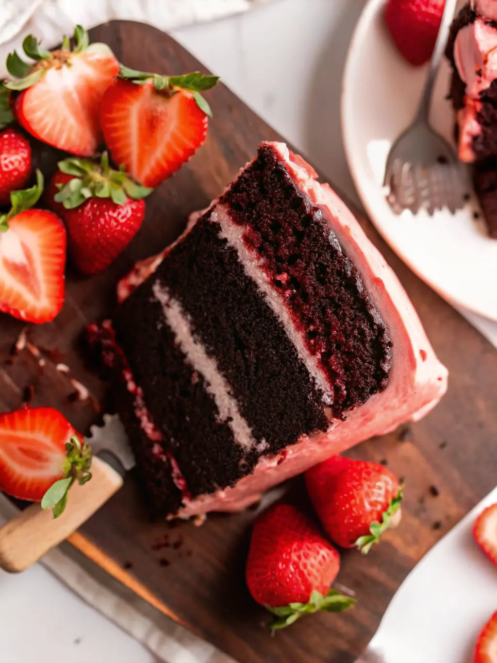 A close-up shot of the Chocolate Covered Strawberry Cake in the process of being assembled: a dark chocolate cake layer being frosted with vibrant pink strawberry buttercream, with more cake layers and a piping bag visible in the soft-focused background. The scene is on a wooden cutting board on a marble countertop with natural light and warm tones. (3:4 ratio).