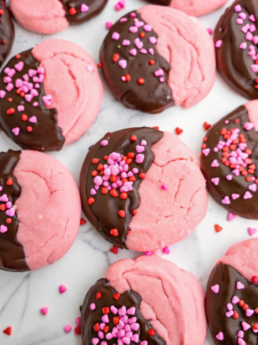 A minimalist flat lay of the key ingredients for pink, crinkly Chocolate Covered Strawberry Cookies on a light grey marble countertop: a bowl of pink cookie dough, a small bowl of dark chocolate chips, and a scattering of red, pink, and white heart sprinkles. A wooden cutting board is partially visible, with soft natural morning light and subtle shadows. Fresh herbs are in the background. NO HANDS OR PEOPLE. 3:4 aspect ratio.