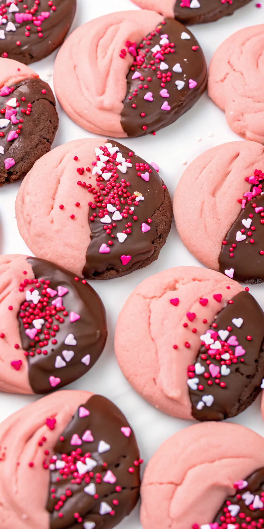 A stack of two perfectly baked, pink, crinkly Chocolate Covered Strawberry Cookies, half-dipped in dark chocolate with heart sprinkles, on a small minimalist white ceramic plate. The cookies showcase their soft texture and glossy chocolate. The setting is a light grey marble countertop with gentle morning light, a wooden cutting board, and fresh herbs blurred in the background. NO HANDS OR PEOPLE. 3:4 aspect ratio.
