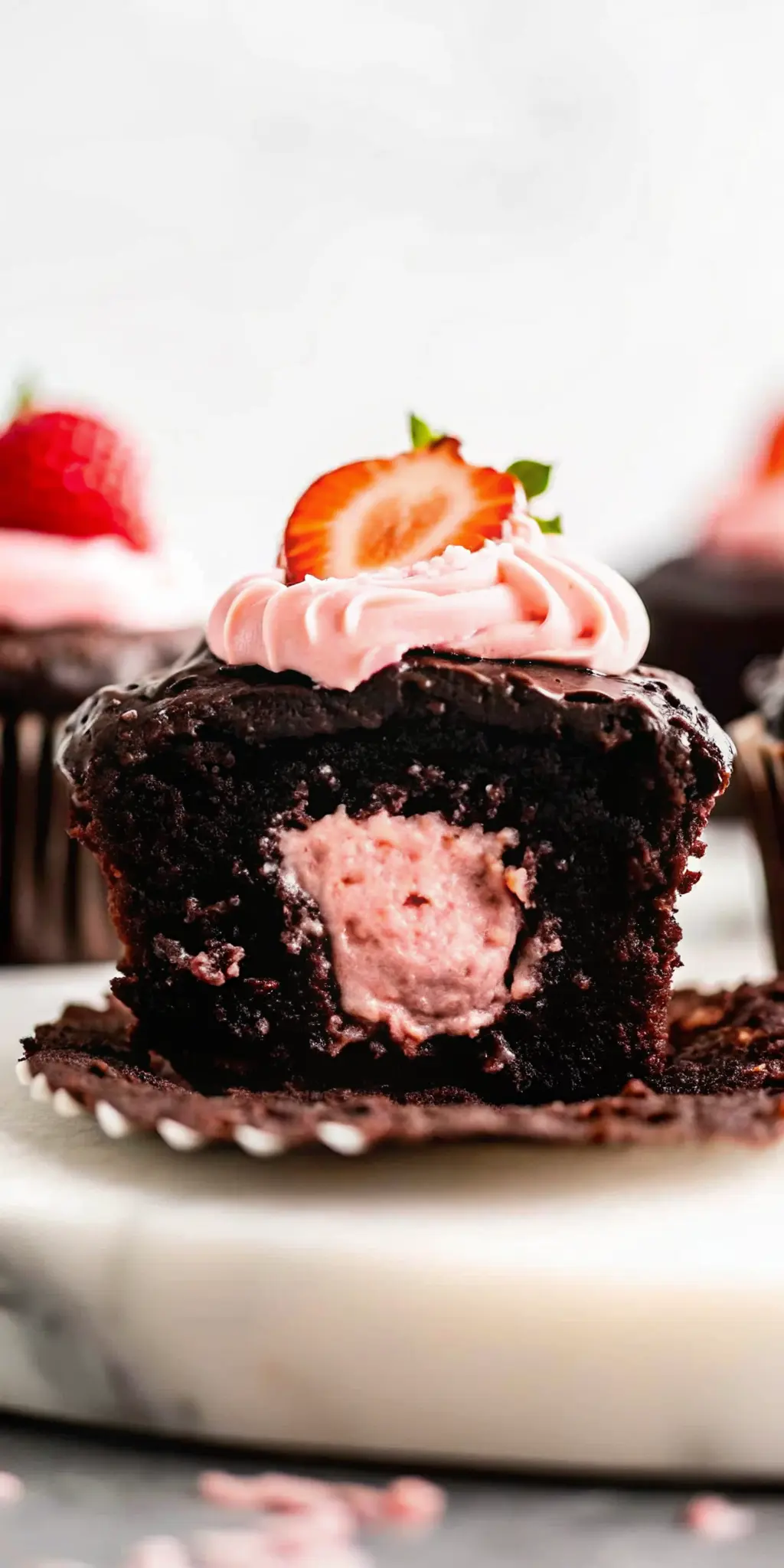 A perfectly plated Chocolate Covered Strawberry Cupcake, showcasing its moist chocolate cake texture, the vibrant pink strawberry heart filling, and the glistening chocolate ganache with a fresh strawberry on top. It's presented on a minimalist white plate on a marble countertop, with soft, warm natural morning light and gentle shadows. (3:4 ratio)