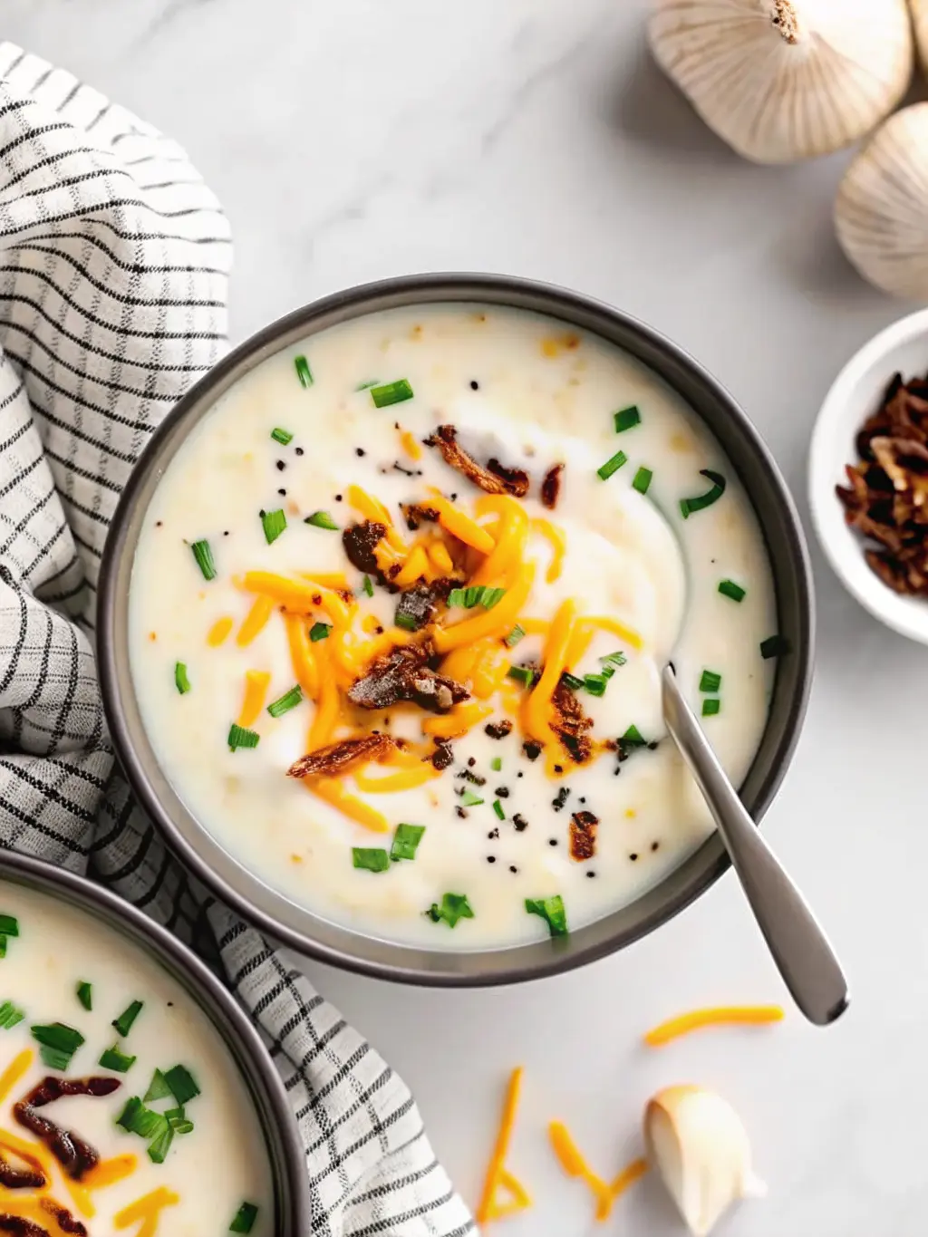 A beautifully styled close-up (3:4 ratio) of a single black ceramic bowl of Creamy Vegan Loaded Baked Potato Soup, showcasing its rich, velvety texture. The soup is adorned with vibrant green chopped chives, perfectly melted shredded orange vegan cheddar, dark brown crispy caramelized onions, and a swirl of white vegan sour cream. A silver spoon is poised for a spoonful. The bowl is on a white marble countertop with soft, warm natural light and delicate shadows. No hands visible.