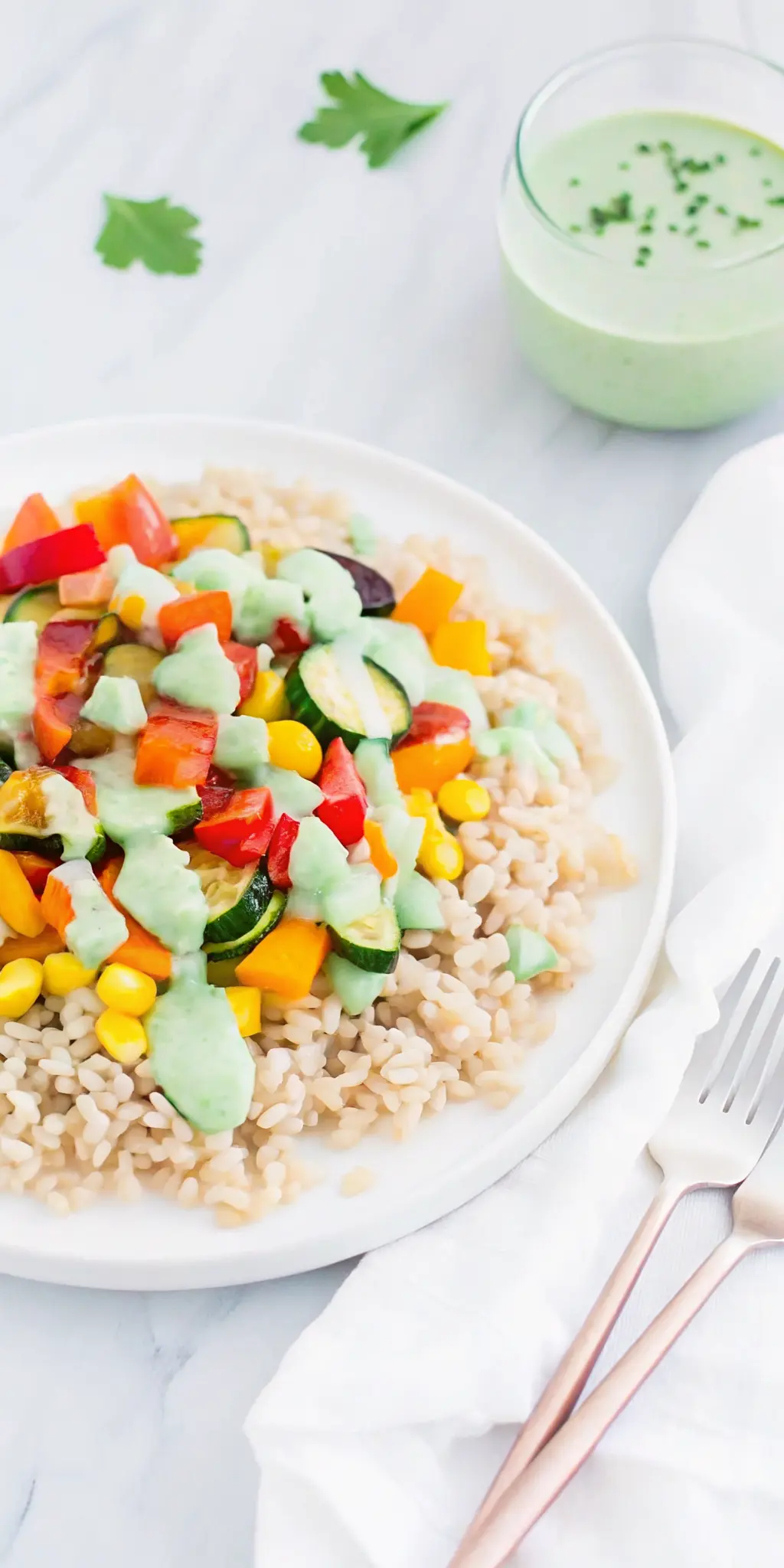 A close-up, slightly overhead shot of a white plate holding a serving of brown rice and colorful roasted vegetables, with the thick, creamy, light green Green Onion Cashew Sauce generously drizzled over the top, highlighting the sauce's texture and sheen. The scene is on a white marble countertop, with a sprig of fresh herbs in the soft-focused background, bathed in natural light.