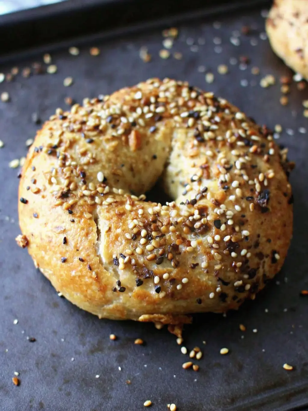 Four unbaked Greek yogurt bagels, shaped into rings, brushed with egg wash and lightly sprinkled with poppy seeds and small, light-colored seeds, lying on parchment paper on a dark baking sheet. The scene is set on a marble countertop under natural morning light, with warm tones and soft shadows. A wooden rolling pin is gently placed nearby. NO HANDS OR PEOPLE. (3:4 ratio)