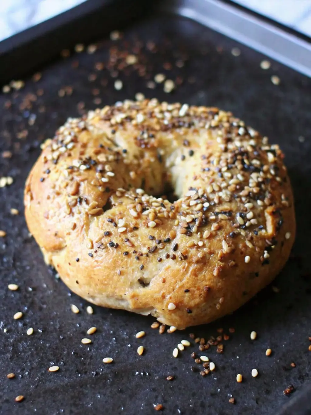 A close-up shot of a single baked Greek yogurt bagel, split open and lightly toasted, revealing its soft, airy, light interior texture. It's adorned with a generous spread of cream cheese and a dusting of the poppy seeds and small, light-colored seeds. The bagel rests on a minimalist white plate on a wooden cutting board, with fresh herbs in the background, bathed in natural morning light with soft shadows and warm tones. NO HANDS OR PEOPLE. (3:4 ratio)