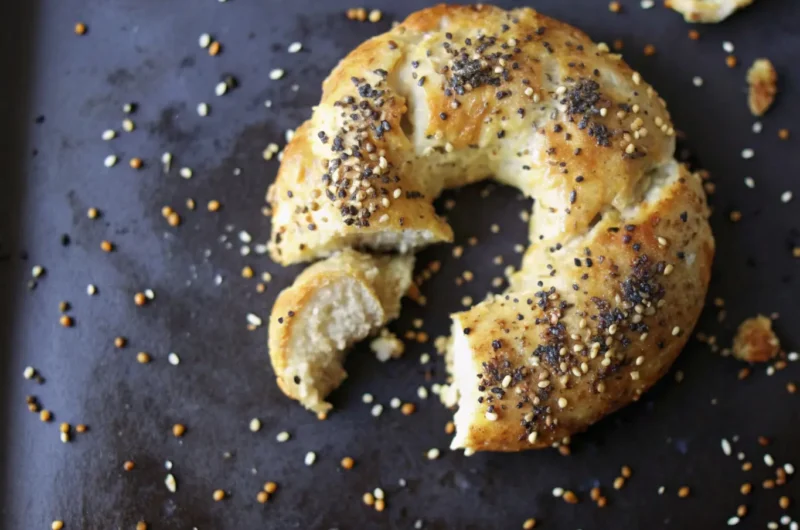 A golden-brown homemade Greek yogurt bagel, torn in half to reveal its soft, chewy, light interior, generously topped with poppy seeds and small, light-colored seeds, resting on a dark baking sheet with some scattered seeds and caramelized drips. The scene is bathed in natural morning light from an east window, highlighting soft shadows. The setting is a clean, tidy marble countertop, with subtle wood accents and a sprig of fresh herbs blurred in the background, warm tones dominant. NO HANDS OR PEOPLE. (4:3 ratio)