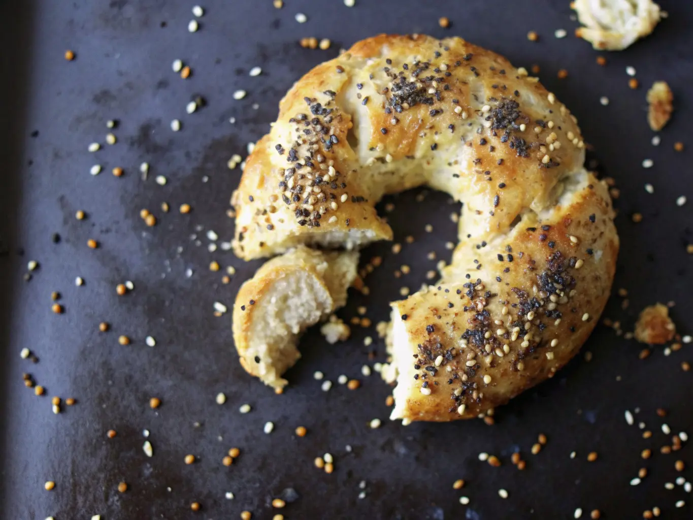 A golden-brown homemade Greek yogurt bagel, torn in half to reveal its soft, chewy, light interior, generously topped with poppy seeds and small, light-colored seeds, resting on a dark baking sheet with some scattered seeds and caramelized drips. The scene is bathed in natural morning light from an east window, highlighting soft shadows. The setting is a clean, tidy marble countertop, with subtle wood accents and a sprig of fresh herbs blurred in the background, warm tones dominant. NO HANDS OR PEOPLE. (4:3 ratio)