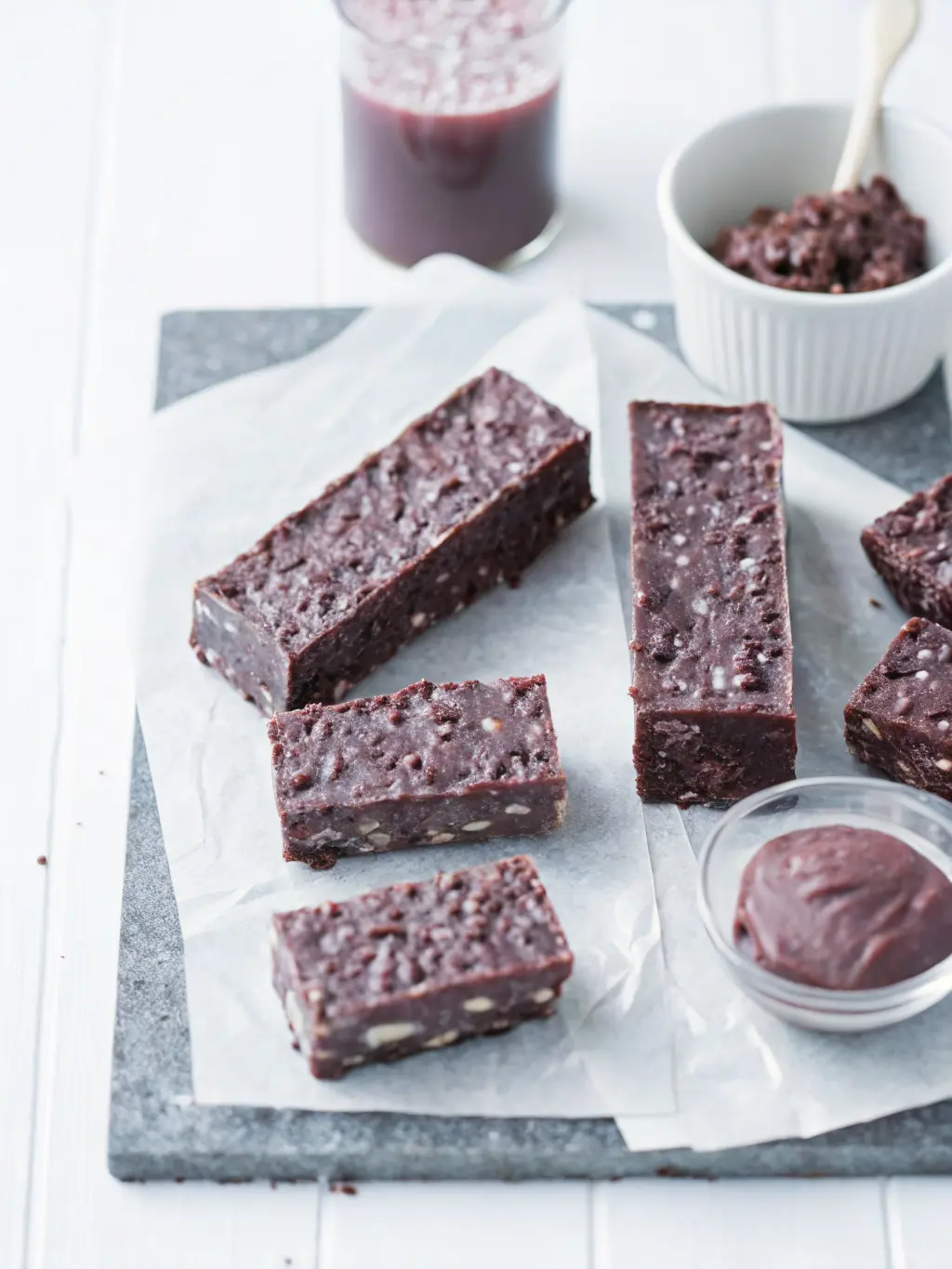 A process shot showing puffed rice cereal being gently folded into a glossy, melted dark chocolate and nut butter mixture in a minimalist white ceramic bowl, using a wooden spatula. The dark wooden cutting board is partially visible in the foreground on a marble countertop. Natural morning light creates soft shadows, with warm tones, and fresh herbs subtly in the background. 3:4 ratio. NO HANDS OR PEOPLE.