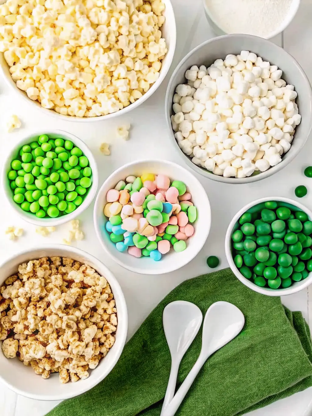 Close-up (3:4 ratio) shot of the ingredients for Loaded Leprechaun Popcorn laid out artfully on a light wooden cutting board, resting on a white marble countertop. Ingredients include a small pile of freshly popped white popcorn, a small bowl of white chocolate wafers, a tiny dish of vanilla extract, a colorful mound of separated Lucky Charms marshmallows (pink, blue, yellow, green shapes), and a scattering of green M&Ms. The scene is illuminated by soft natural morning light, with fresh green herbs subtly blurred in the background, creating a warm and tidy presentation.