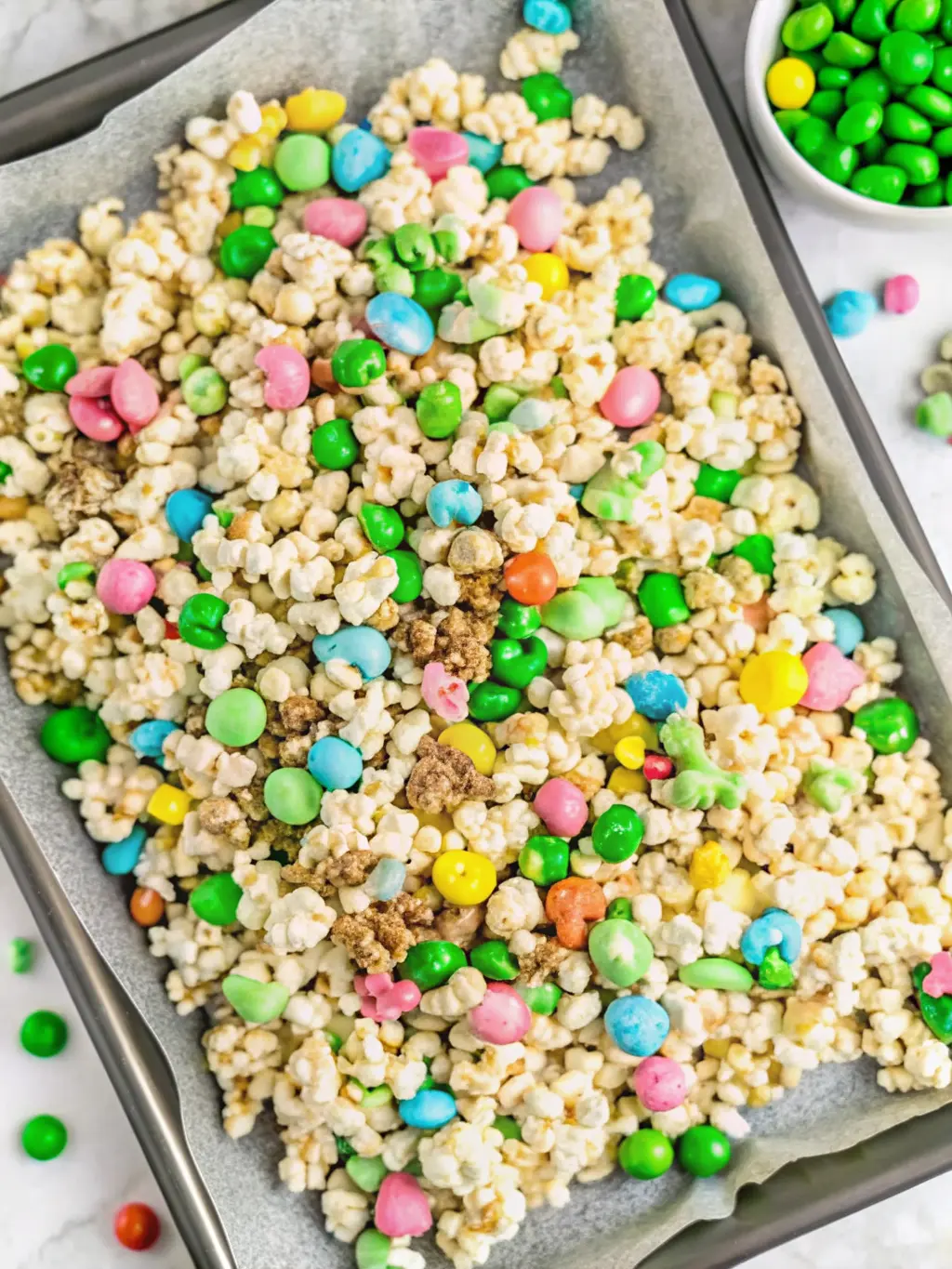 A process-focused shot (3:4 ratio) depicting the moment fluffy white popcorn is being gently tossed with glossy melted white chocolate and vibrant, colorful Lucky Charms marshmallows in a large, textured white ceramic mixing bowl. The popcorn is partially coated, showcasing the transition. The bowl sits on a white marble countertop, with warm, soft shadows and natural morning light highlighting the textures. The wooden cutting board is subtly visible nearby, ensuring consistency with the kitchen's visual identity.