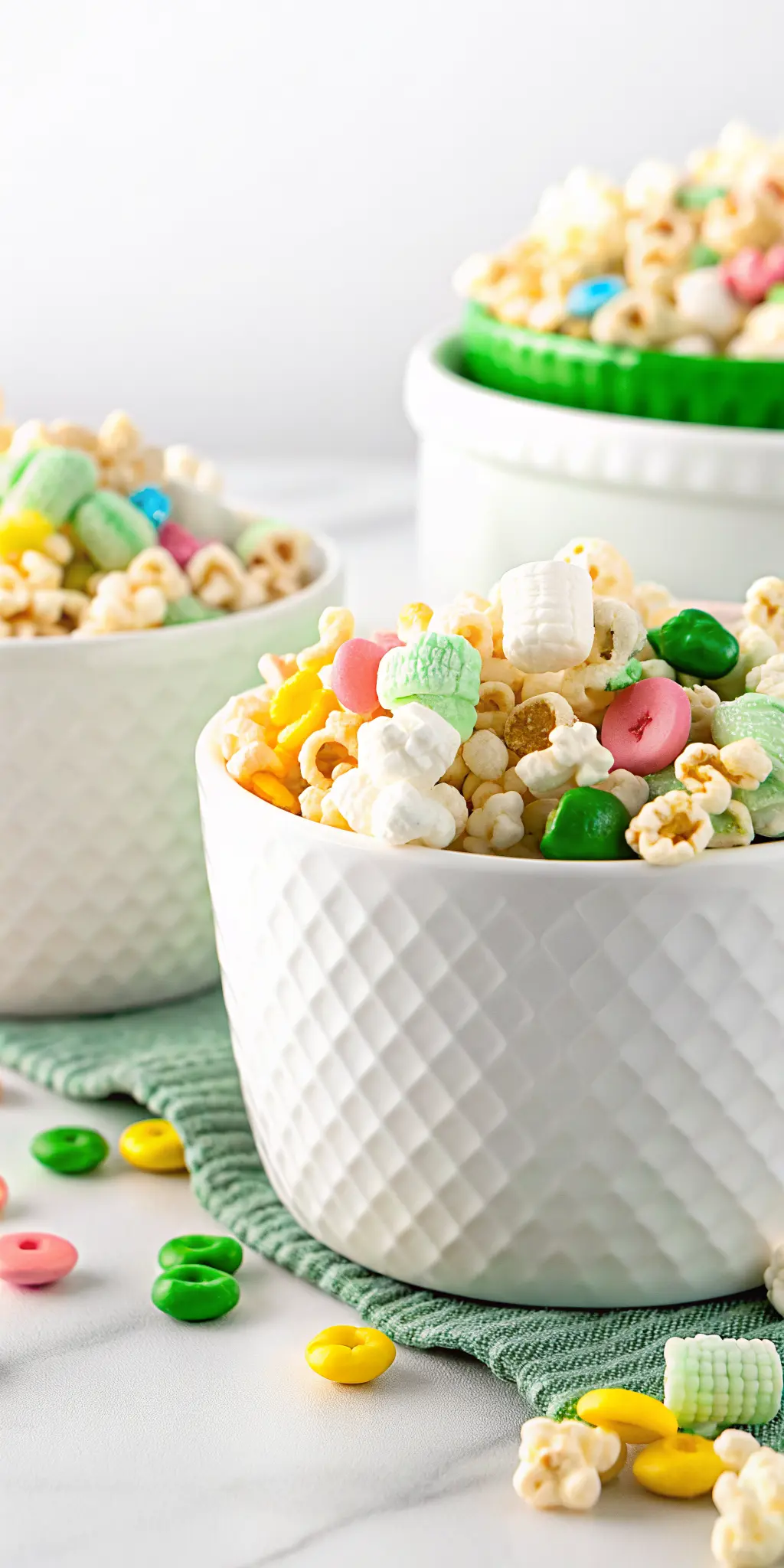 An artistic serving shot (3:4 ratio) of Loaded Leprechaun Popcorn presented in a beautiful, textured white ceramic bowl, showcasing the crisp popcorn, the even white chocolate coating, and the vivid, distinct shapes and colors of the Lucky Charms marshmallows and green candies. The bowl is positioned slightly off-center on a pristine white marble countertop. A sprig of fresh green herbs is subtly visible in the soft background, adding a touch of natural elegance. Soft morning light creates a warm and inviting atmosphere, highlighting the texture and appeal of the snack.