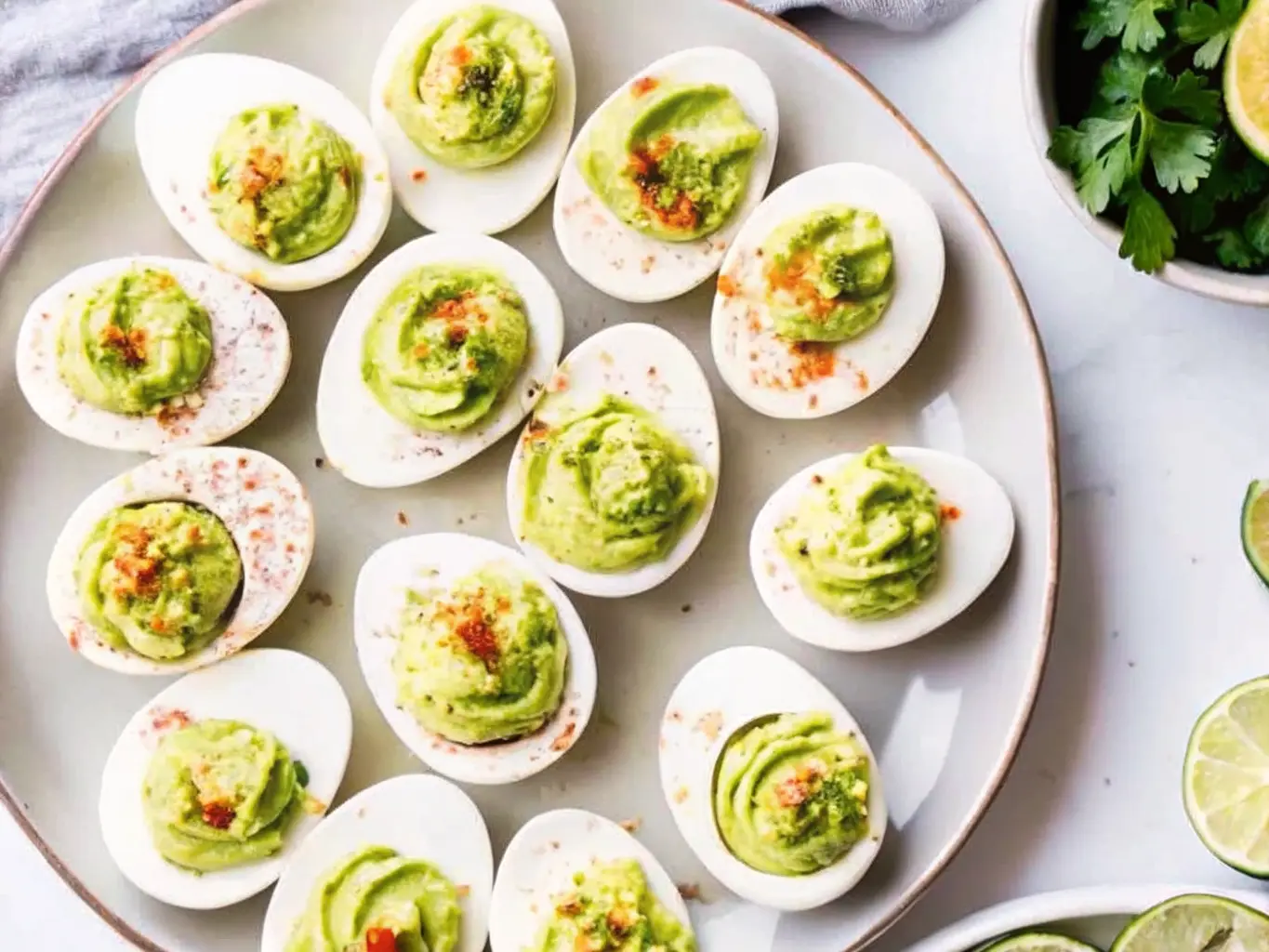 A 4:3 ratio hero shot of approximately 12 perfectly piped deviled egg halves arranged on a light blue ceramic platter, garnished generously with smoked paprika and finely chopped fresh chives. The background is softly blurred, showing a small ceramic bowl of paprika and another small ceramic bowl of chopped chives on a marble countertop. The scene is illuminated by natural morning light from an east window, casting soft shadows. The overall tone is warm, clean, and tidy, embodying a lived-in kitchen aesthetic. NO HANDS OR PEOPLE.