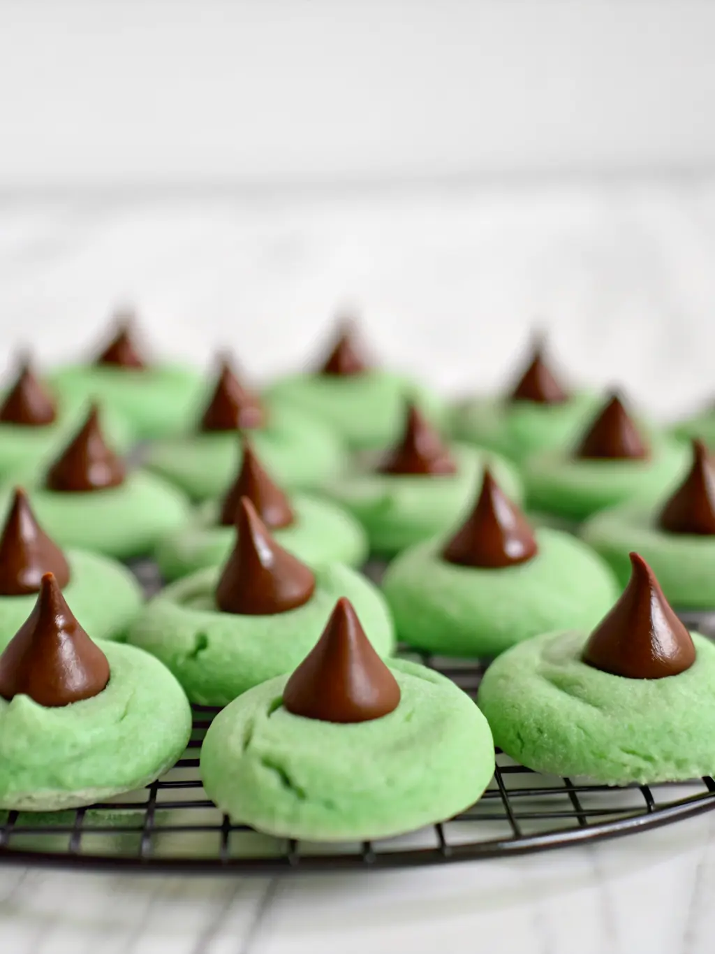 A minimalist flat lay showcasing neatly organized ingredients for Mint Chocolate Kiss Blossom Cookies: bowls of flour, granulated sugar, softened butter, mint extract in a small bottle, green gel food coloring, and a pile of unwrapped chocolate kisses on marble countertops. The same wooden cutting board is subtly visible in the background, with a sprig of fresh mint. Natural morning light, soft shadows, warm tones, clean and tidy, no hands, 3:4 aspect ratio.