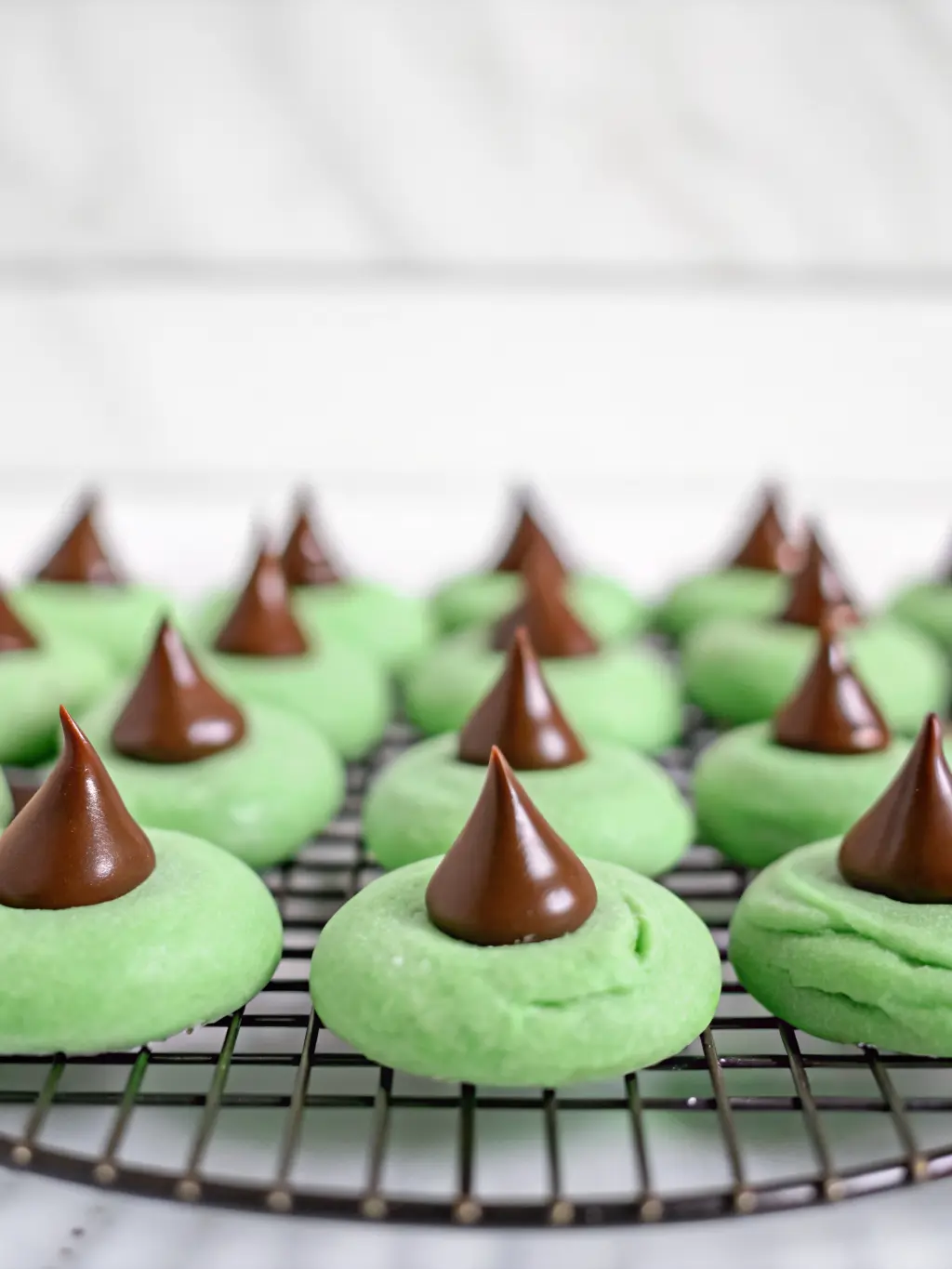 A baking sheet filled with perfectly round, unbaked green cookie dough balls, some already rolled in granulated sugar and gently indented, ready for chocolate kisses. The cookies are arranged on parchment paper on a sturdy baking sheet resting on the familiar wooden cutting board, with a hint of marble countertop. Natural morning light, soft shadows, warm tones, clean and tidy, no hands, 3:4 aspect ratio.