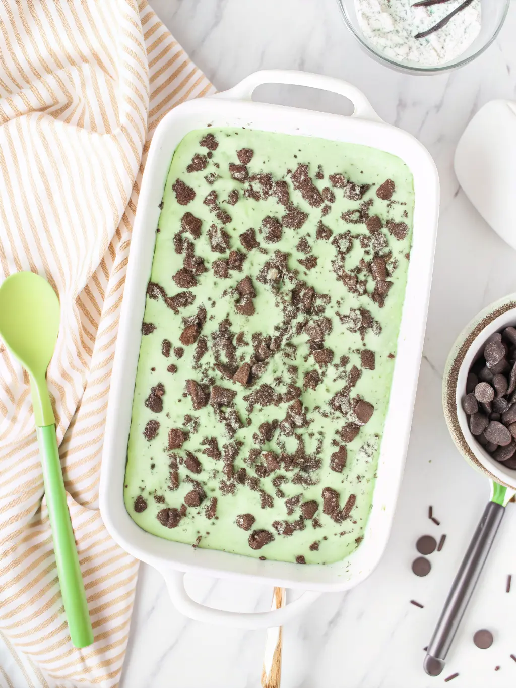A collection of ingredients for No-Bake Shamrock Lasagna laid out on a marble countertop: packages of Oreo cookies, a block of cream cheese, a tub of whipped topping, a box of instant pistachio pudding mix, green food coloring, a bowl of melted butter, and a small dish of maraschino cherries. All are arranged neatly around a ceramic bowl and a wooden cutting board, under natural morning light with soft shadows and warm tones. NO HANDS OR PEOPLE. 3:4 ratio.