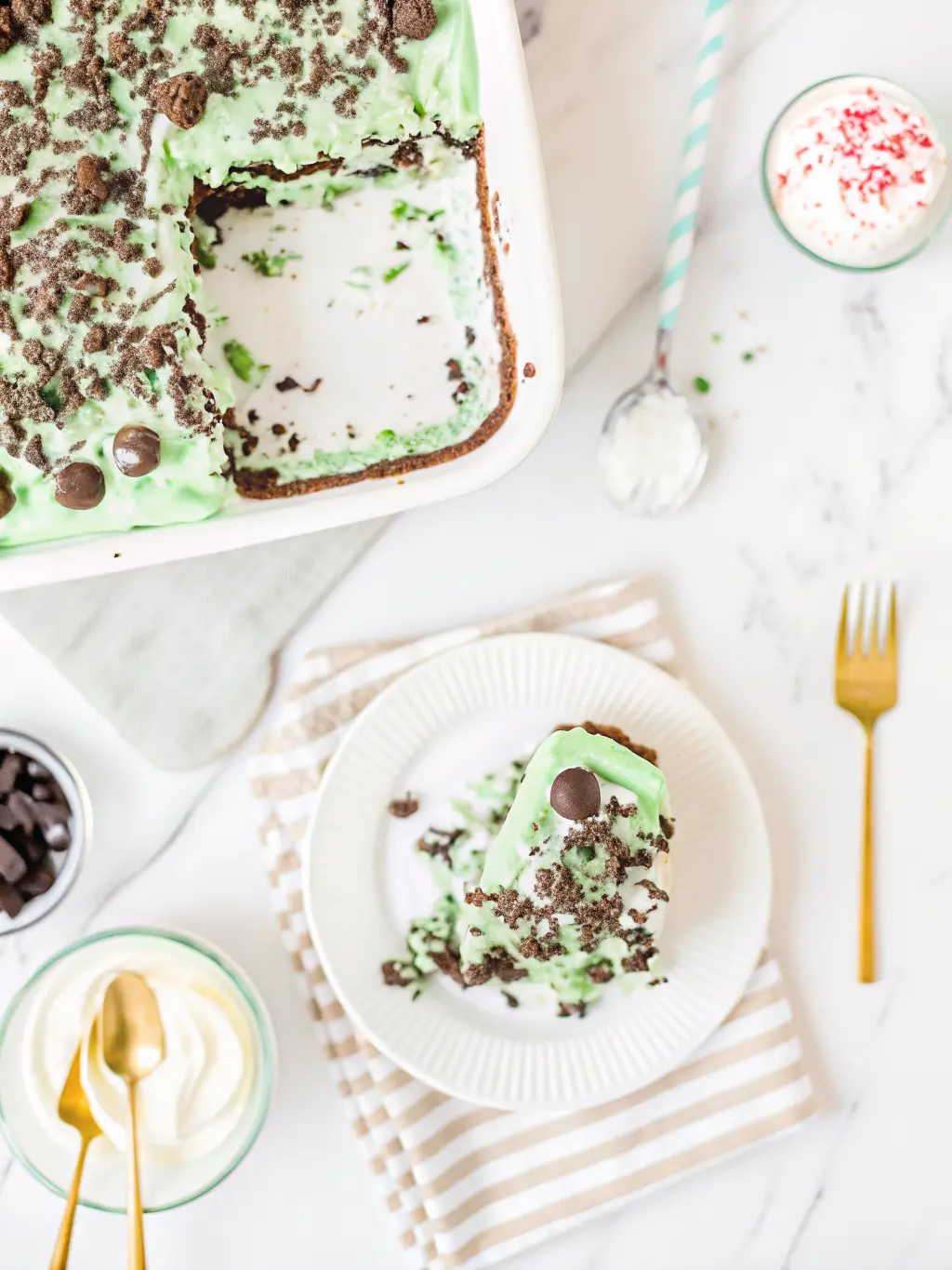 A process shot showing the layers of the Shamrock Lasagna being assembled in a clear 9x13 inch baking dish. The dark Oreo crust is visible at the bottom, topped by the creamy white layer, with the light green mint layer being gently spread on top with an offset spatula. The setting is a clean marble countertop with soft natural morning light and subtle wood accents in the background. NO HANDS OR PEOPLE. 3:4 ratio.