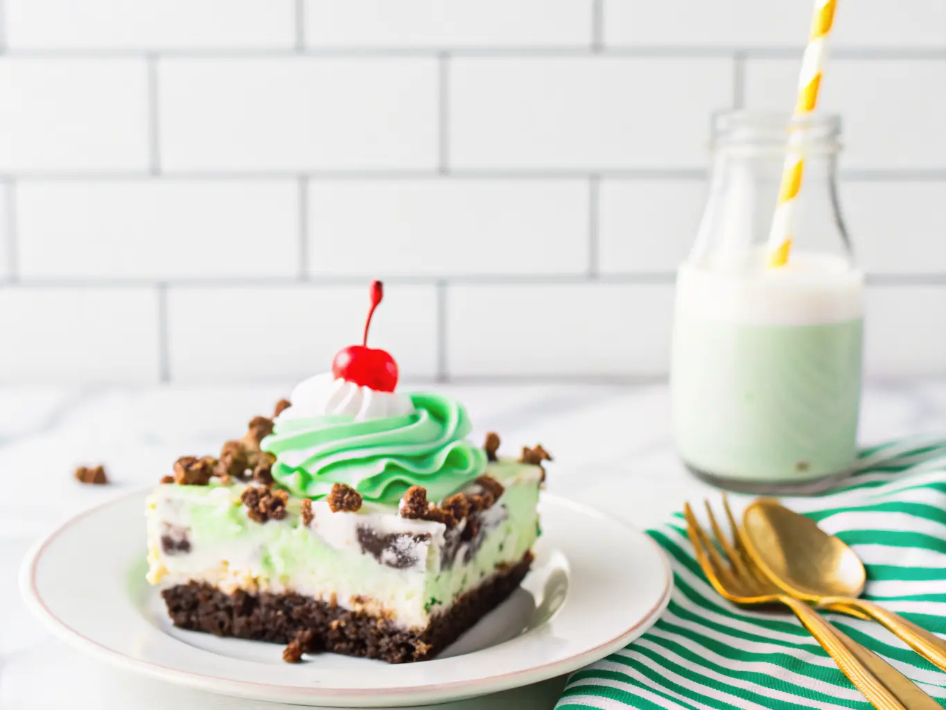 A generous slice of No-Bake Shamrock Lasagna dessert, showcasing its distinct layers: dark Oreo crust, creamy white filling, and vibrant mint green top layer. It's elegantly topped with a swirl of whipped cream, crushed Oreo pieces, sparkling green sprinkles, and a bright red maraschino cherry. The slice rests on a minimalist white plate with a gold fork nearby, on a yellow and white striped linen. A small glass bottle of milk with a blue and white striped straw is in the background, against a white subway tile backsplash, all bathed in natural morning light on a marble countertop with subtle wood accents. Soft shadows, warm tones, clean and tidy presentation. 4:3 ratio. NO HANDS OR PEOPLE.