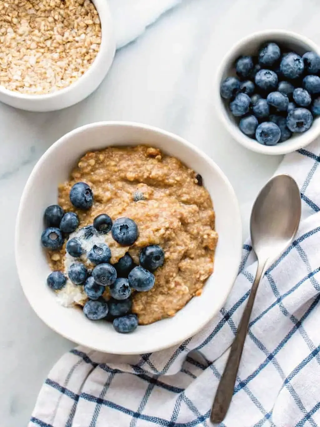 A beautifully arranged flat lay (3:4 ratio) of raw quinoa grains in a small white ceramic pinch bowl, a carton of oat milk, a jar of almond butter, and a scattering of fresh, plump blueberries on a light wooden cutting board. All on a white marble countertop, with natural morning light and soft shadows. A sprig of fresh mint in the background. Clean and tidy. NO HANDS OR PEOPLE.