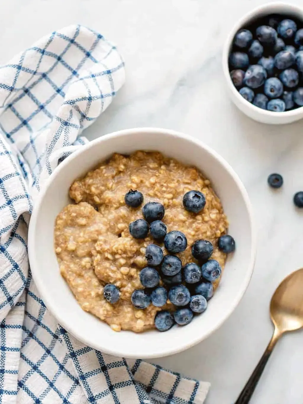 A minimalist white ceramic saucepan (3:4 ratio) containing fluffy cooked quinoa, gently steaming, with a wooden spoon just resting inside. The saucepan is on a clean white marble countertop, with a subtle wood accent (edge of a cutting board) in the background. Natural morning light creates warm tones and soft shadows. Clean and tidy. NO HANDS OR PEOPLE.