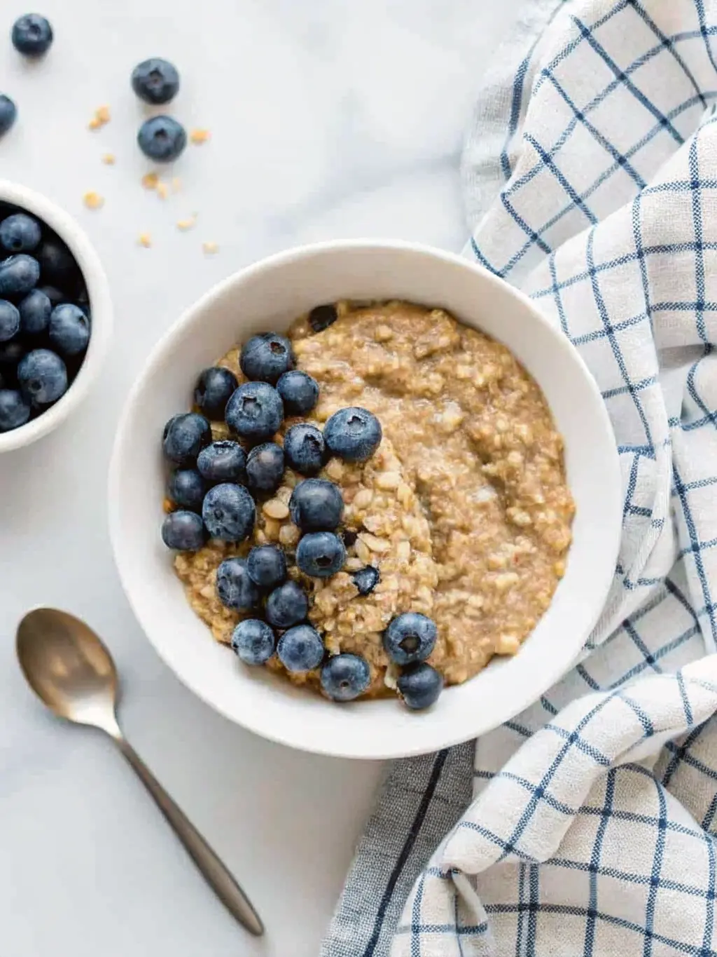 A close-up, slightly overhead shot (3:4 ratio) of a minimalist white ceramic bowl of the finished vegan quinoa breakfast bowl, showcasing the fluffy texture of the quinoa, the burst of blue from the berries, and the smooth, creamy consistency of the almond butter drizzle with visible hemp seeds. The bowl is centered on a white marble countertop, illuminated by natural morning light, with soft shadows. A blue and white checkered linen peeks from the side. Clean and tidy. NO HANDS OR PEOPLE.