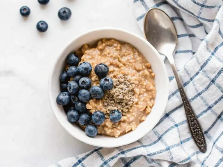 A minimalist white ceramic bowl (4:3 ratio) filled with a warm, fluffy vegan quinoa breakfast bowl, generously topped with fresh, vibrant blueberries, a luscious drizzle of creamy almond butter, and a sprinkle of light green hemp seeds. The bowl sits on a white marble countertop, bathed in natural morning light from an east window, casting soft shadows. A blue and white checkered linen napkin is neatly folded beside it, with a vintage-style silver spoon resting on the right. A hint of a wooden cutting board is in the background, along with a sprig of fresh mint. Warm tones, clean and tidy presentation. NO HANDS OR PEOPLE.