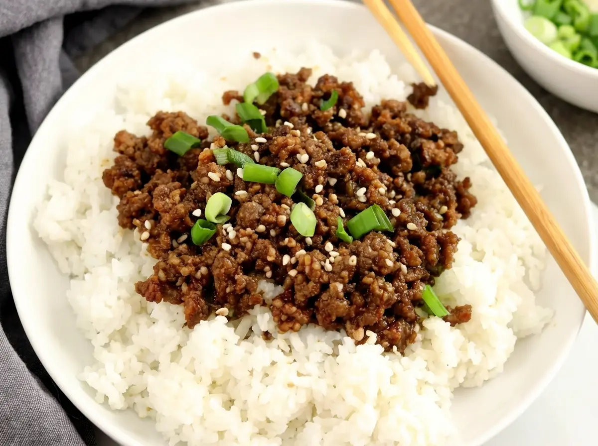 A vibrant Korean Ground Beef Bowl, presented in a minimalist white ceramic bowl on a light marble countertop. The bowl is filled with fluffy white rice, topped generously with savory, dark brown ground beef, scattered with bright green sliced green onions and white sesame seeds. Wooden chopsticks rest on the rim of the bowl. Soft natural morning light from an east window illuminates the scene, creating warm tones and gentle shadows, with a grey linen napkin and a small bowl of fresh green onions blurred in the background, all clean and tidy. (4:3 aspect ratio)
