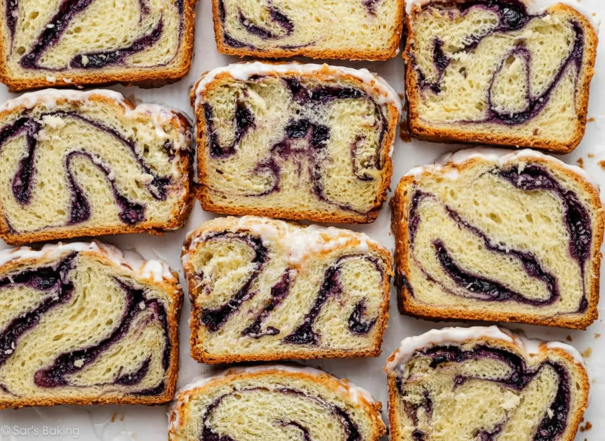 A top-down shot (4:3 ratio) of several perfectly baked slices of Lemon Blueberry Babka, showcasing the intricate purple blueberry swirls and a glistening white lemon glaze. The slices are arranged neatly on a minimalist white plate on a marble countertop, with hints of a wooden cutting board in the soft background. Natural morning light streams from an east window, creating soft shadows and warm tones. Clean and tidy presentation, no hands or people.