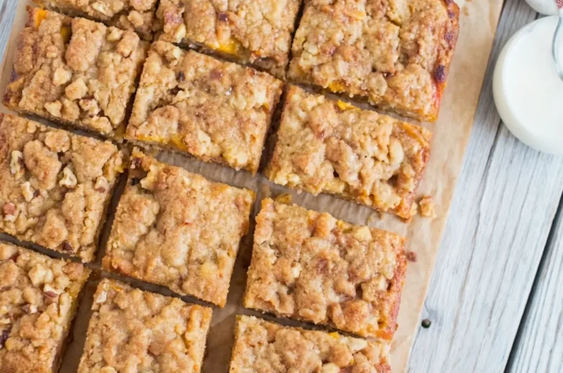 A hero shot (4:3 ratio) of several freshly baked Peaches and Cream Crumble Bars cut into squares on a light wooden cutting board. One bar is slightly pulled forward, showcasing the golden-brown crumble topping, the soft, juicy peach layer, and the creamy white base. In the softly focused background, a light blue enamel mug filled with dark coffee and a folded white kitchen towel with subtle blue stripes are visible. The scene is illuminated by natural morning light from an east window, casting soft shadows on the marble countertop. The presentation is clean and tidy with warm tones.