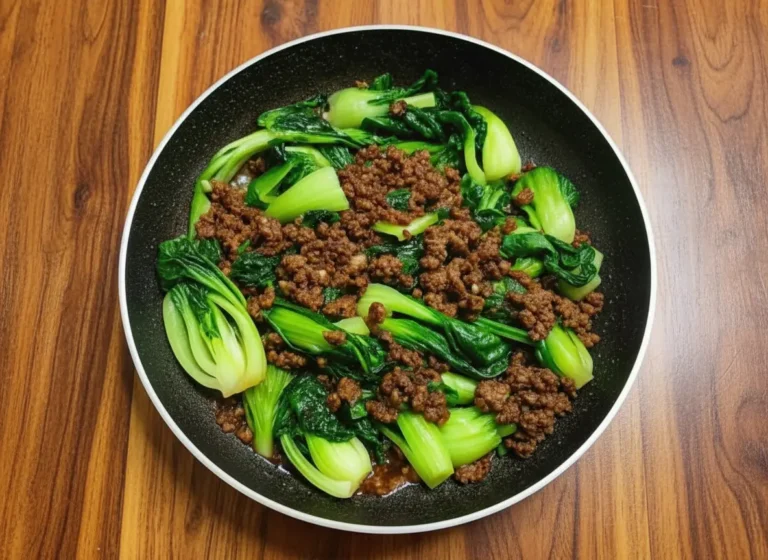 A high-angle hero shot of a dark skillet centered on a marble countertop with wood accents. The skillet contains Stir-Fried Bok Choy With Beef, featuring browned ground beef crumbles and vibrant green baby bok choy halves coated in a glossy dark brown sauce. Chopped green onions are scattered on top. Natural morning light casts soft shadows from the east window. Fresh herbs are blurred in the background.