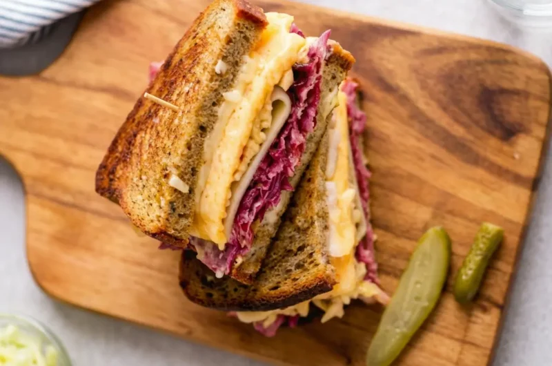 A beautifully stacked Reuben sandwich, cut in half, on toasted rye bread, showcasing distinct layers of thinly sliced corned beef, tangy sauerkraut, perfectly melted Swiss cheese, and a hint of creamy Russian dressing. Two crisp dill pickle spears rest on the same rustic wooden cutting board. Another similarly prepared Reuben sandwich is artfully blurred in the background. The scene is set on a light marble countertop, bathed in natural morning light from an east window, with soft shadows and warm tones, presented cleanly and tidily. Fresh herbs are subtly visible in the soft-focus background. (4:3 ratio)