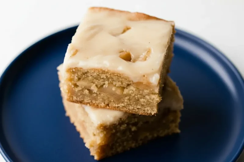 A hero shot of two stacked Brown Butter Apple Blondies, square-shaped, light golden brown with a creamy off-white glaze slightly dripping, showcasing their soft, chewy texture and visible apple pieces. The blondies are on a minimalist white plate, placed on a light marble countertop with warm natural morning light from an east window. Soft shadows, a hint of fresh green apple or herbs in the very soft-focused background, and a clean, tidy presentation. (4:3 ratio)