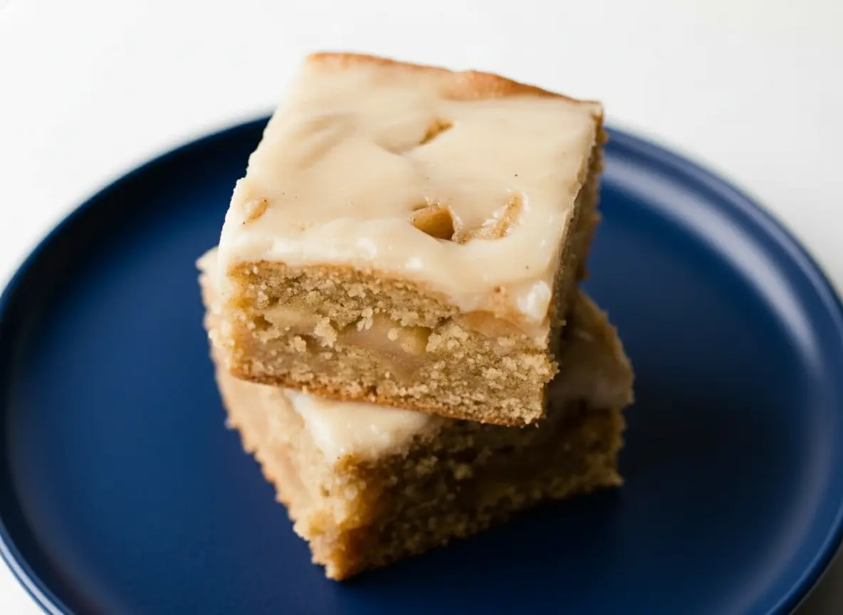 A hero shot of two stacked Brown Butter Apple Blondies, square-shaped, light golden brown with a creamy off-white glaze slightly dripping, showcasing their soft, chewy texture and visible apple pieces. The blondies are on a minimalist white plate, placed on a light marble countertop with warm natural morning light from an east window. Soft shadows, a hint of fresh green apple or herbs in the very soft-focused background, and a clean, tidy presentation. (4:3 ratio)