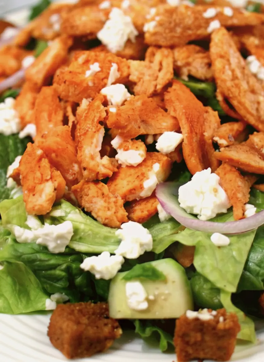 A clean and organized flat lay of all raw ingredients for Buffalo Chicken Caesar Salad on a light wooden cutting board. This includes a head of fresh romaine lettuce, an un-sliced red onion, a whole English cucumber, boneless chicken breasts, a bowl of buffalo sauce, a block of feta cheese, and a bottle of Caesar dressing. The items are neatly arranged on a white marble countertop under soft natural morning light, with fresh herbs subtly in the background.