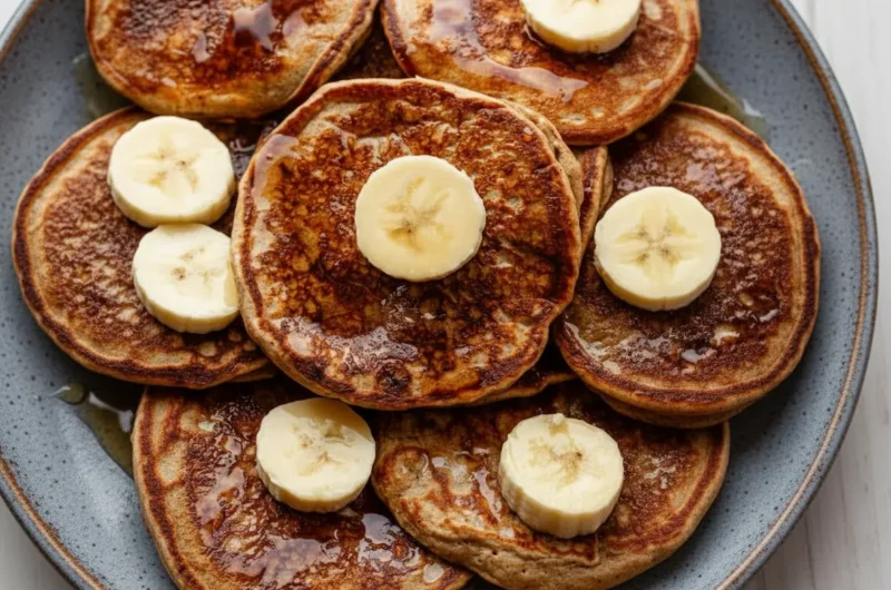 A stack of golden brown healthy banana pancakes, generously drizzled with pure maple syrup, topped with fresh banana slices on a minimalist white ceramic plate. The plate sits on a light grey marble countertop with a subtle wood accent in the background, illuminated by soft natural morning light from an east window. The focus is on the rich, textured surface of the pancakes and the glistening syrup, with a clean and tidy presentation. (4:3 ratio)
