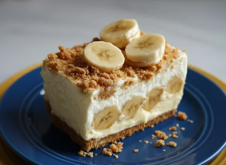 A perfect slice of No-Bake Banana Pudding Cheesecake, showcasing its distinct layers of golden graham cracker crust, creamy white filling with visible banana slices, and a generous topping of whipped cream, more banana slices, and crumbled graham cracker crumbs. The dessert is on a minimalist white plate on a marble countertop with natural morning light creating soft shadows and warm tones. A wooden cutting board and fresh herbs are subtly blurred in the background, creating a clean and tidy presentation. (4:3 ratio)