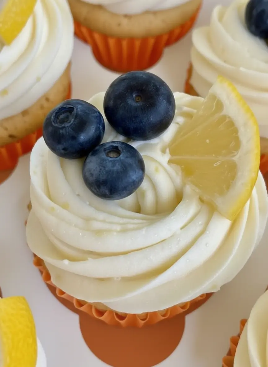 A clean composition of fresh ingredients for Blueberry Lemon Cupcakes on a wooden cutting board: whole lemons, a small ceramic bowl brimming with vibrant fresh blueberries, a stick of butter, and a measuring cup of flour, all under natural morning light on a marble countertop with soft shadows. (3:4 ratio)