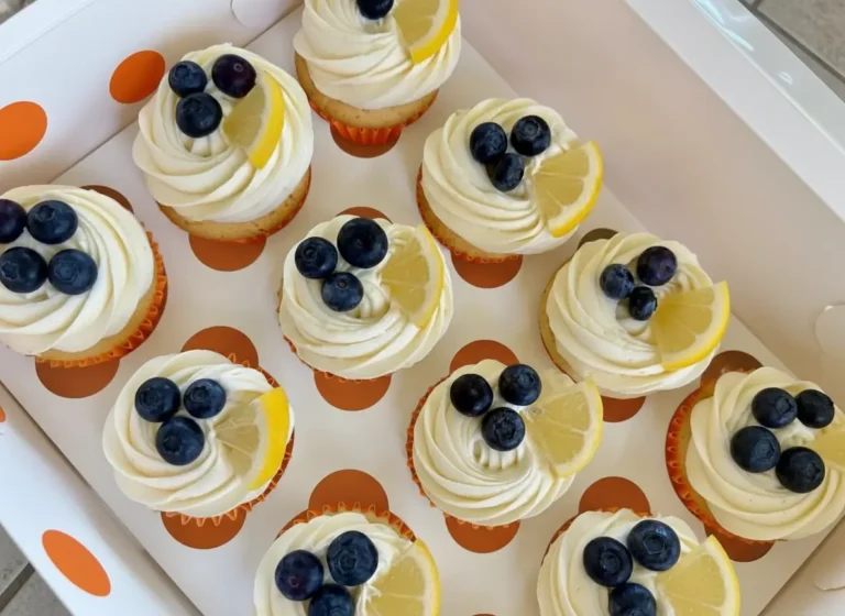 A hero shot of several Blueberry Lemon Cupcakes displayed in a white cupcake box with brown polka dots. Each cupcake has perfectly swirled white frosting, topped with fresh, plump blueberries and a delicate wedge of lemon. The scene is bathed in natural morning light on a marble countertop, with subtle wood accents in the soft-focused background, clean and tidy presentation. (4:3 ratio)