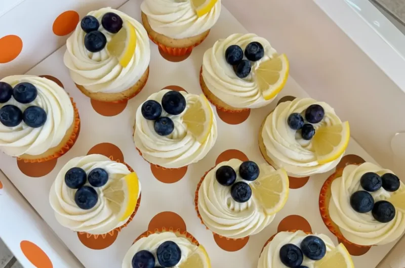 A hero shot of several Blueberry Lemon Cupcakes displayed in a white cupcake box with brown polka dots. Each cupcake has perfectly swirled white frosting, topped with fresh, plump blueberries and a delicate wedge of lemon. The scene is bathed in natural morning light on a marble countertop, with subtle wood accents in the soft-focused background, clean and tidy presentation. (4:3 ratio)