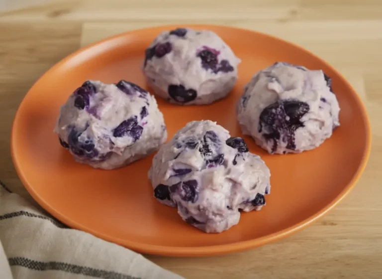 A close-up, high-angle shot of four pale violet blueberry protein cheesecake bites, shaped like perfect rounded scoops and studded with whole dark blueberries, sitting elegantly on a minimalist white ceramic plate. The plate rests on a dark wooden cutting board over a clean marble countertop. Warm, soft morning light streams from an east window, casting gentle shadows across the scene with a hint of fresh green herbs in the soft-focus background.