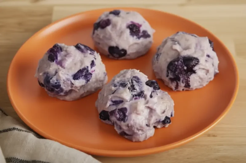 A close-up, high-angle shot of four pale violet blueberry protein cheesecake bites, shaped like perfect rounded scoops and studded with whole dark blueberries, sitting elegantly on a minimalist white ceramic plate. The plate rests on a dark wooden cutting board over a clean marble countertop. Warm, soft morning light streams from an east window, casting gentle shadows across the scene with a hint of fresh green herbs in the soft-focus background.