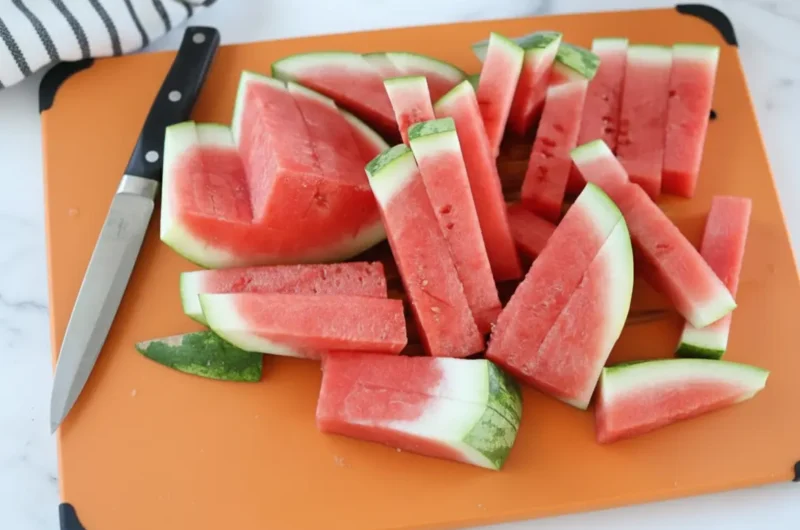 A white minimalist plate overflowing with perfectly cut, vibrant red watermelon sticks, each displaying a thin strip of white and green rind. Fresh mint leaves are scattered around the base of the sticks. The scene is bathed in natural morning light from an east window, highlighting the marble countertop with a subtle wood accent in the background. Soft shadows, warm tones, and a clean, tidy presentation. (4:3)