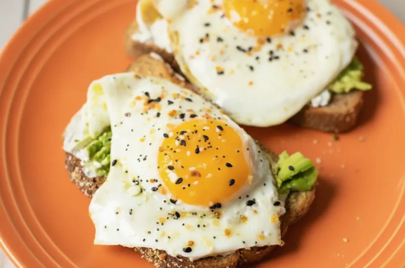 A high-angle 4:3 shot of two slices of 10-Minute Avocado Egg Toast on a minimalist white ceramic plate. The toast is dark, seedy multigrain bread topped with thick mashed green avocado and two perfectly fried sunny-side-up eggs with bright orange runny yolks. The eggs are generously sprinkled with black and white everything bagel seasoning and red pepper flakes. The plate sits on a white marble countertop with a soft morning light coming from the side, creating gentle shadows. A small sprig of fresh cilantro is visible in the soft-focus background.