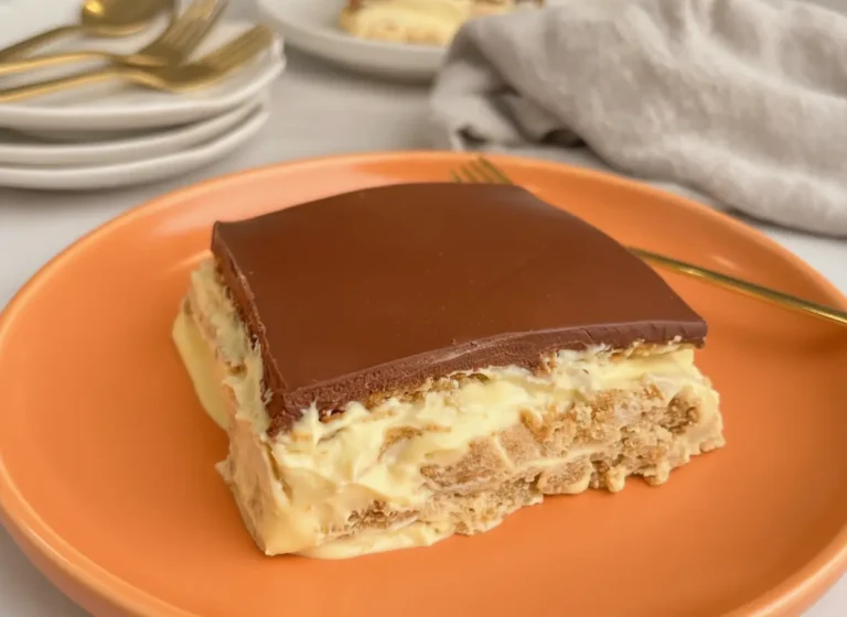 A hero shot of a single square slice of No-Bake Eclair Cake served on a minimalist white ceramic plate. The cake shows distinct layers of softened golden-brown graham crackers and thick, pale yellow vanilla cream. The top is covered in a smooth, dark brown, semi-matte chocolate glaze. A gold fork rests beside the plate on a white marble countertop. Natural morning light creates soft shadows. In the background, a soft grey linen napkin and a hint of a wooden cutting board are visible. No hands, no people.
