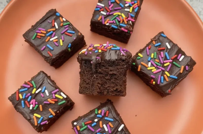 A top-down hero shot of sixteen perfectly square Chocolate Donut-Frosted Cake Bites on a minimalist white ceramic plate. The bites feature a dark, moist crumb topped with a thick, glossy dark chocolate frosting and a dense layer of rainbow cylinder sprinkles. The plate sits on a white marble countertop with a wooden cutting board partially visible. Soft morning light from an east window creates gentle shadows and warm tones.
