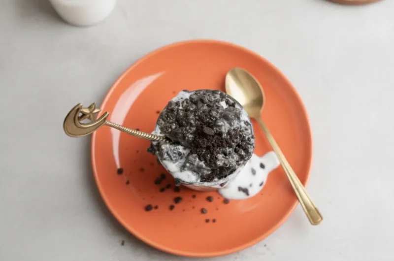 A glass jar of Cookies & Cream Overnight Oats with dramatic black cocoa swirls and white creamy oat layers, topped with crushed cookie bits. A decorative gold crescent moon and star spoon is tucked into the jar. The jar sits on a round wooden tray on a marble countertop in bright morning light. Minimalist white background with soft shadows.