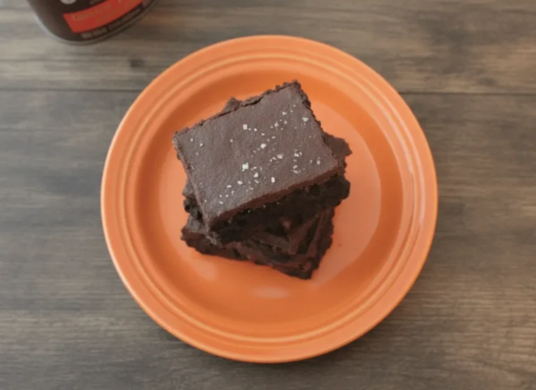 A hero shot of a towering stack of three thick, square Dark Chocolate Sea Salt Brownies on a minimalist white ceramic plate. The brownies are a deep, dark ebony color with a dense, fudgy texture visible on the cut sides. The top of each brownie is covered in a thick, smooth dark chocolate ganache frosting, generously sprinkled with large, translucent flakes of coarse sea salt. The scene is set on a white marble countertop with a wooden cutting board nearby. Soft, warm morning light streams in from an east-facing window, creating gentle shadows. A small sprig of fresh mint is visible in the soft-focus background.