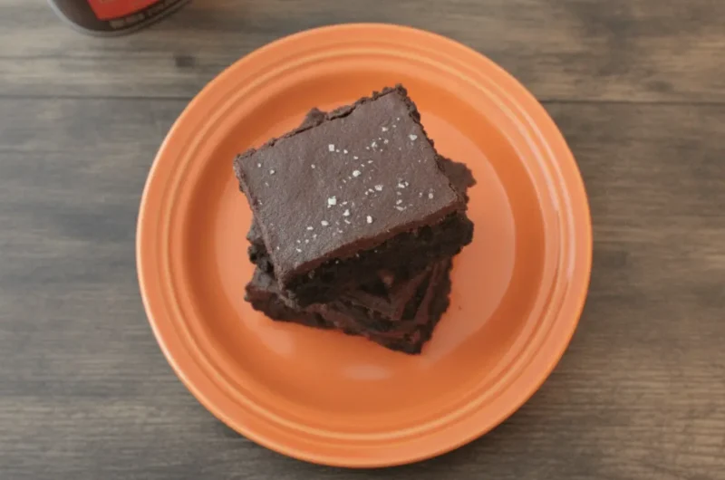 A hero shot of a towering stack of three thick, square Dark Chocolate Sea Salt Brownies on a minimalist white ceramic plate. The brownies are a deep, dark ebony color with a dense, fudgy texture visible on the cut sides. The top of each brownie is covered in a thick, smooth dark chocolate ganache frosting, generously sprinkled with large, translucent flakes of coarse sea salt. The scene is set on a white marble countertop with a wooden cutting board nearby. Soft, warm morning light streams in from an east-facing window, creating gentle shadows. A small sprig of fresh mint is visible in the soft-focus background.